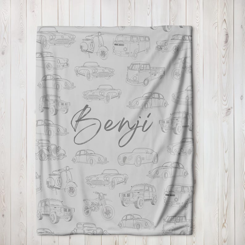 May include: A light gray blanket featuring a pattern of vintage cars, scooters, and vans in a line drawing style. The name "Benji" is written in a cursive font across the center of the blanket.