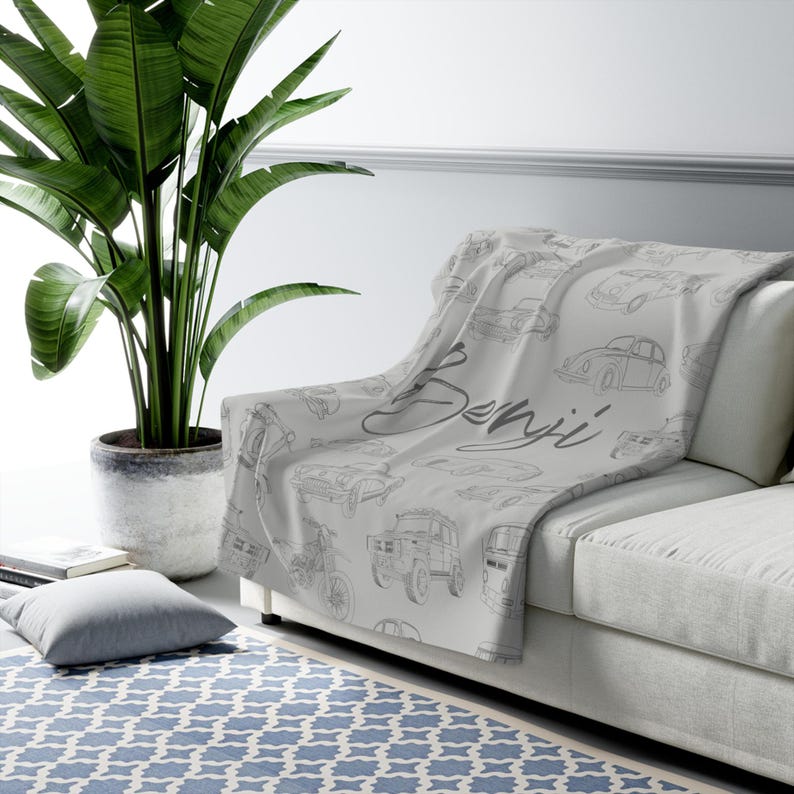 May include: A light gray throw blanket draped over a white sofa. The blanket features a repeating pattern of sketched cars in gray and the name "Benji" in cursive. A large green plant in a gray pot is to the left.