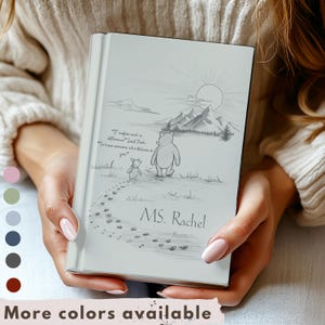 May include: A personalized notebook with a pencil sketch of Winnie the Pooh and Piglet walking towards mountains and a sun. The notebook is inscribed with the name "MS. Rachel" and a quote. The words "More colors available" are at the bottom.