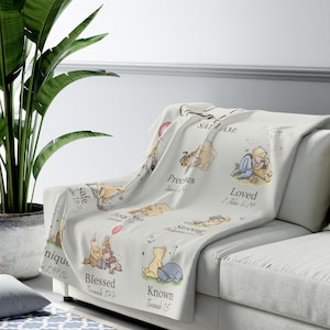 May include: A light-coloured throw blanket draped over a sofa, featuring illustrations of Winnie the Pooh characters. The blanket has text that reads "Safe", "Precious", "Loved", "Unique", "Blessed", "Known", and "Strong".