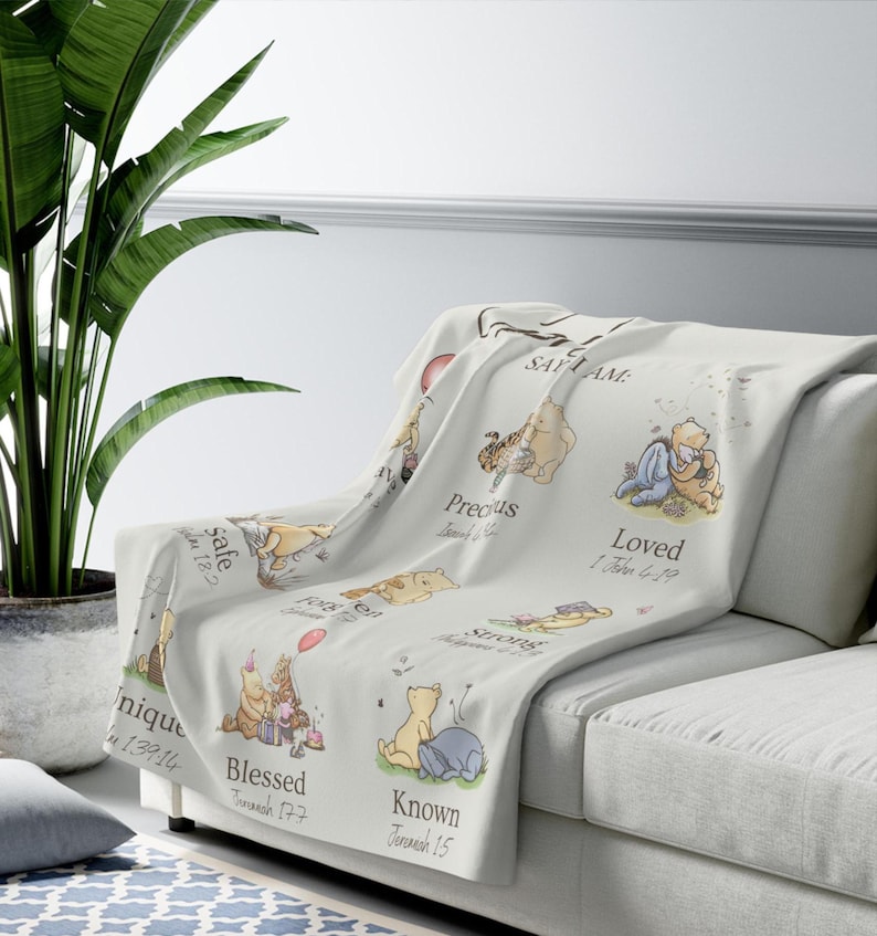 May include: A light-coloured throw blanket draped over a sofa, featuring illustrations of Winnie the Pooh characters. The blanket has text that reads "Safe", "Precious", "Loved", "Unique", "Blessed", "Known", and "Strong".