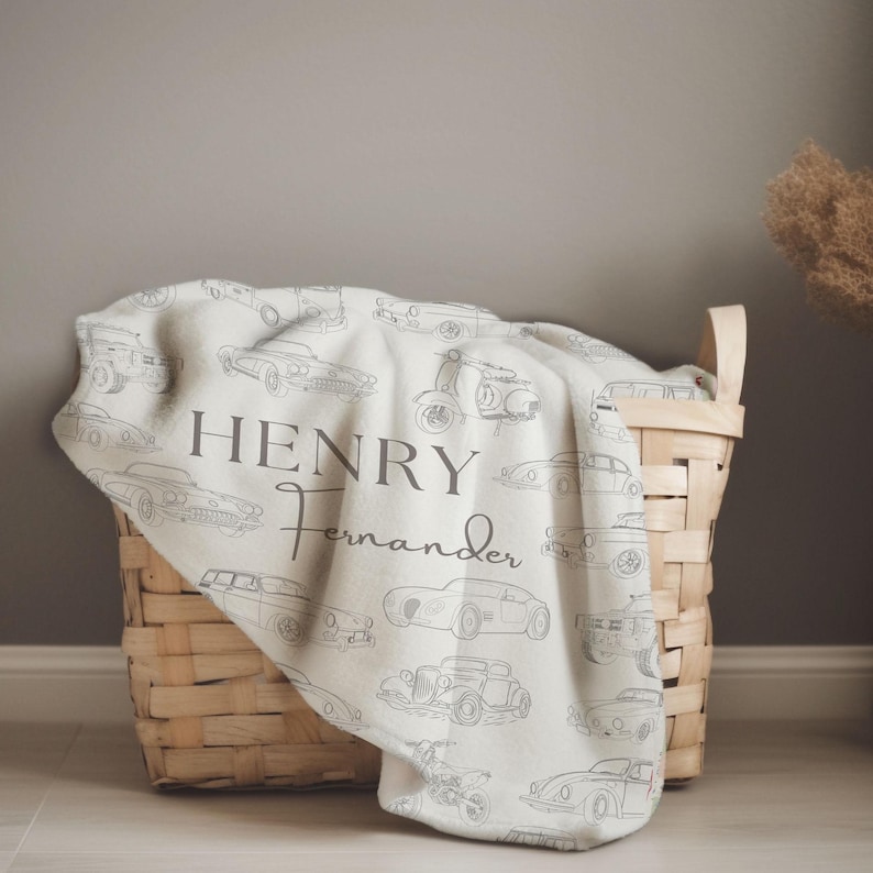May include: A cream-colored blanket with a pattern of vintage cars and scooters. The blanket is draped over a woven basket. The name "HENRY FERNANDER" is printed in gray script on the blanket.