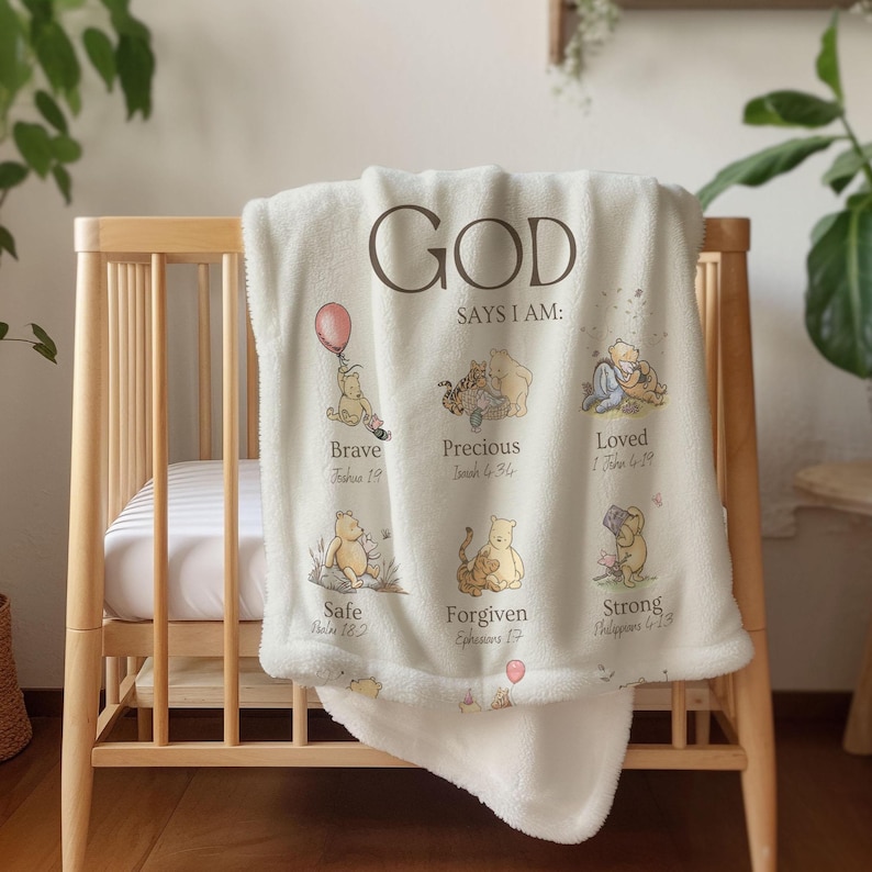 May include: Cream baby blanket with the text "GOD SAYS I AM" and illustrations of a bear. The blanket features the words "Brave", "Precious", "Loved", "Safe", "Forgiven", and "Strong". The blanket is draped over a wooden cot.