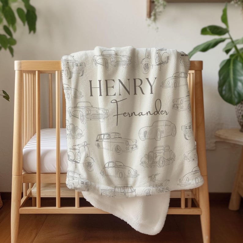 May include: Cream-colored baby blanket with a pattern of vintage cars and scooters in gray. The blanket is draped over a wooden crib. The name "HENRY" and the last name "Fernander" are printed on the blanket in gray.