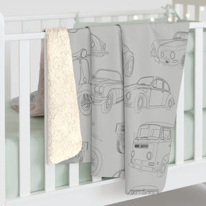 May include: A light gray baby blanket featuring line drawings of vintage cars, scooters, and a van. The blanket is draped over a white crib rail, with a cream-colored sherpa blanket visible on the left side.
