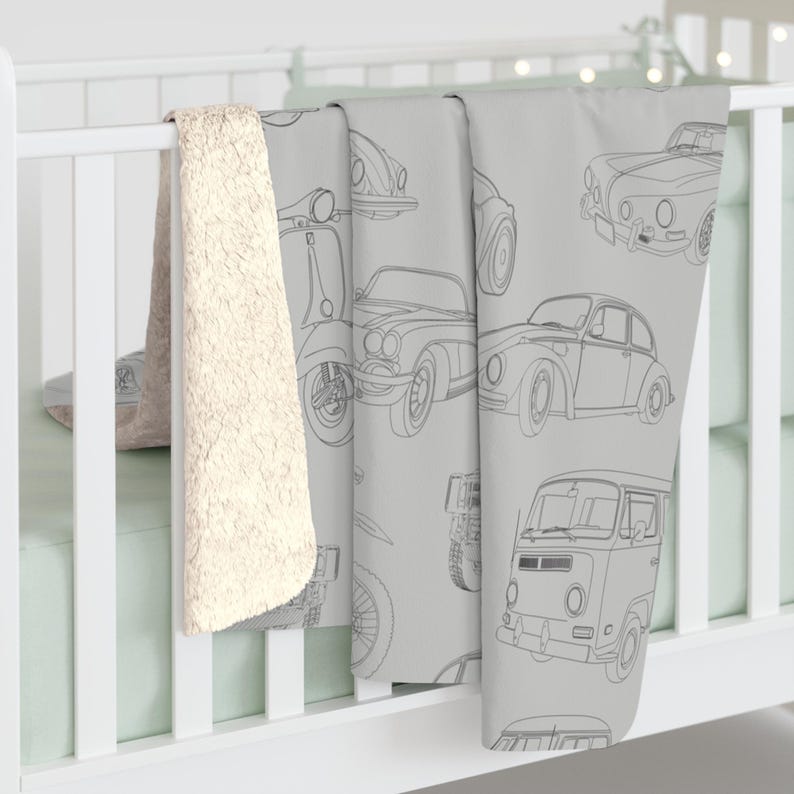 May include: A light gray baby blanket featuring line drawings of vintage cars, scooters, and a van. The blanket is draped over a white crib rail, with a cream-colored sherpa blanket visible on the left side.