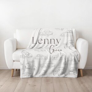 May include: A white throw blanket draped over a white sofa, featuring a pattern of vintage car sketches in gray. The blanket has the name "Lenny Green" printed in gray script.