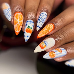 Op de afbeelding: Close-up van een hand met lange, puntige nagels met ingewikkelde nail art. Ontwerpen omvatten sinaasappelschijfjes, een ruitpatroon met een sinaasappel en blauw-witte patronen met vissen en een potje sinaasappelsap.