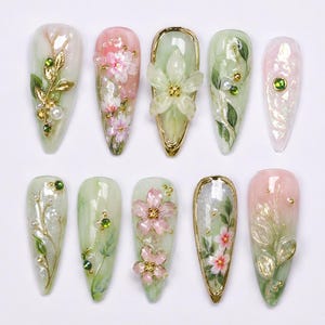 Könnte beinhalten: Ein Set aus zehn mandelförmigen Press-on-Nägeln mit botanischem Motiv in zarten Rosa- und Salbeigrüntönen. Die Designs zeigen handgemalte Kirschblüten, goldene Blattakzente, kleine weiße Perlen sowie winzige grüne Strasssteine als Verzierungen.