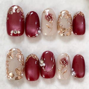 May include: A set of ten artificial nails in various designs. The nails feature a mix of burgundy and nude colors, adorned with gold floral patterns, rhinestones, and pearls. Some nails have a solid burgundy color, while others showcase intricate designs.