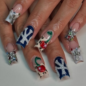Puede incluir: Una manicura con uñas largas y decoradas. Algunas uñas presentan una base blanca con un diseño de cruz plateada, mientras que otras tienen una base azul con un diseño blanco. Otras uñas tienen un diseño de estampado de leopardo con una cereza. También se ve un anillo dorado con perlas.