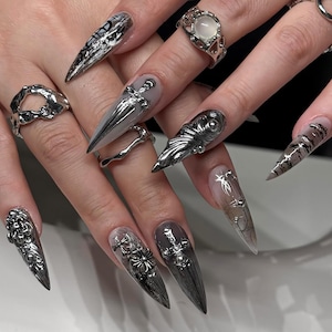 Puede incluir: Primer plano de manos adornadas con uñas largas, puntiagudas, plateadas y negras. Las uñas presentan diseños intrincados, incluyendo adornos y patrones metálicos. También se ven anillos plateados con varios diseños, incluyendo una piedra preciosa.