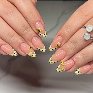 May include: Almond-shaped nails featuring a nude base with yellow French tips decorated with black polka dots. Small, three-dimensional yellow and black bee charms are applied to select nails, creating a textured, playful design on the manicured tips.
