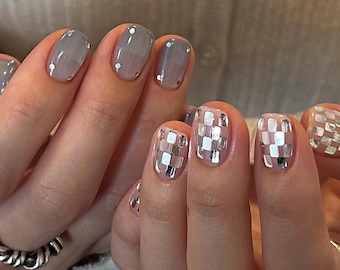 Op de afbeelding: Gemanicureerde vingernagels met een gespleten ontwerp. De linkerhand toont een doorschijnende grijze lak met kleine zilveren accenten aan de basis. De rechterhand heeft een reflecterend zilveren ruitpatroon op een transparante basis. Twee zilveren ringen.