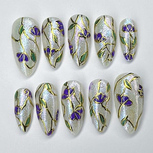 May include: Set of ten artificial nails with a pearlescent finish. Each nail is adorned with a gold outline of flowers, purple petals, and green leaves. The nails are in various sizes and shapes.