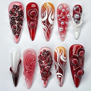 Puede incluir: Una colección de diez uñas artificiales largas en forma de almendra. Las uñas presentan varios diseños, incluyendo remolinos rojos y blancos, acentos de perlas y adornos rojos texturizados. Cada uña tiene un acabado brillante, mostrando intrincado arte de uñas.