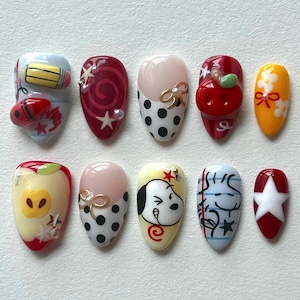 Può includere: Un set di dieci unghie finte con vari disegni. I disegni includono una matita e una coccinella, un vortice rosso, pois, una mela, Snoopy e una stella bianca. I colori includono rosso, bianco, giallo, rosa e blu.