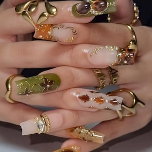May include: Long square-shaped artificial nails featuring a mix of olive green, amber, and clear tones with gold filigree, floral patterns, diamond shapes, and small white pearls. Several gold-toned metal rings are worn on the fingers.