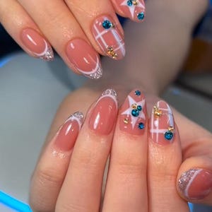 May include: Nude-toned almond-shaped nails featuring silver glitter French tips, white star accents, white grid patterns, and small blue and gold rhinestones embedded on the surface of the nails for a decorative and textured appearance.