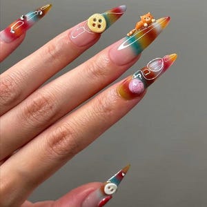 May include: Long, pointed fingernails with a rainbow gradient design. Each nail features a unique 3D embellishment, including a button, a small orange cat, a pair of glasses, and a ball of yarn. The nails are painted with a clear base and a colorful tip.