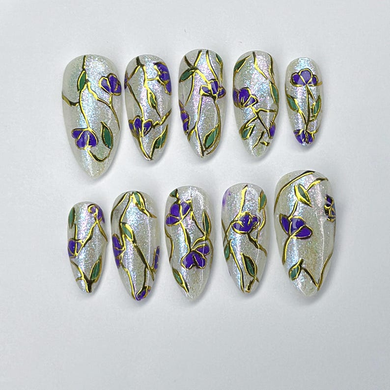 May include: Set of ten artificial nails with a pearlescent finish. Each nail is adorned with a gold outline of flowers, purple petals, and green leaves. The nails are in various sizes and shapes.
