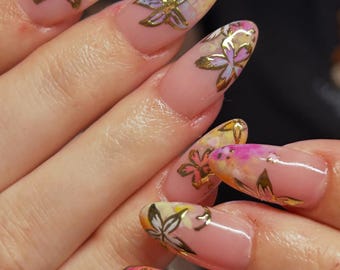 May include: Close-up of manicured nails with a floral design. The nails are painted with a light pink base and feature gold metallic flower accents. The flowers are set against a watercolor-style background with yellow, pink, and blue hues.