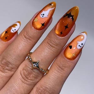 May include: Almond-shaped nails featuring a vibrant orange cat-eye polish base. The design includes white ghost figures, black bats, and small black star accents. A gold ring with a star-shaped center and a second ring with moon and star charms.