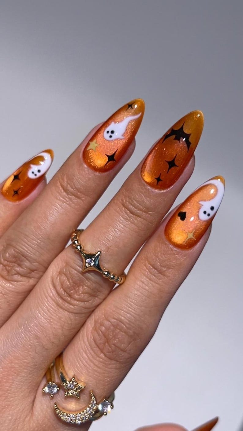 May include: Almond-shaped nails featuring a vibrant orange cat-eye polish base. The design includes white ghost figures, black bats, and small black star accents. A gold ring with a star-shaped center and a second ring with moon and star charms.