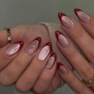 May include: A set of long, almond-shaped nails with a red French tip and a shimmering, light pink base. The nails are well-manicured and polished, showcasing a modern and elegant style. A gold ring with small gemstones is visible.