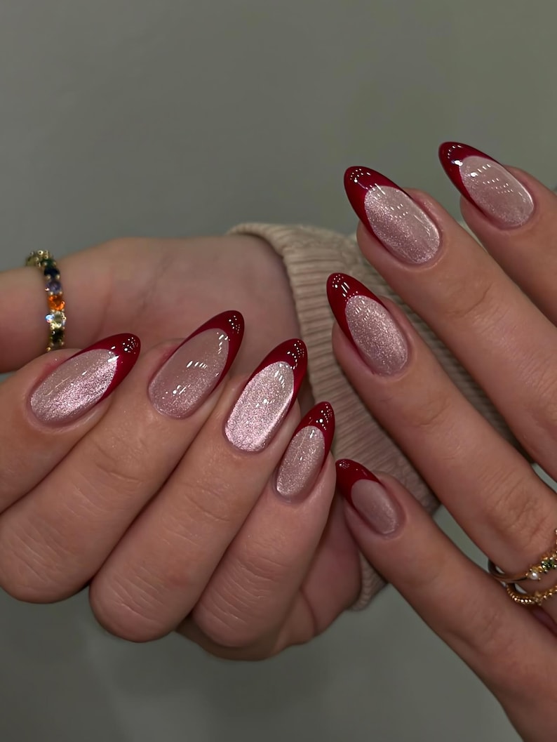 May include: A set of long, almond-shaped nails with a red French tip and a shimmering, light pink base. The nails are well-manicured and polished, showcasing a modern and elegant style. A gold ring with small gemstones is visible.