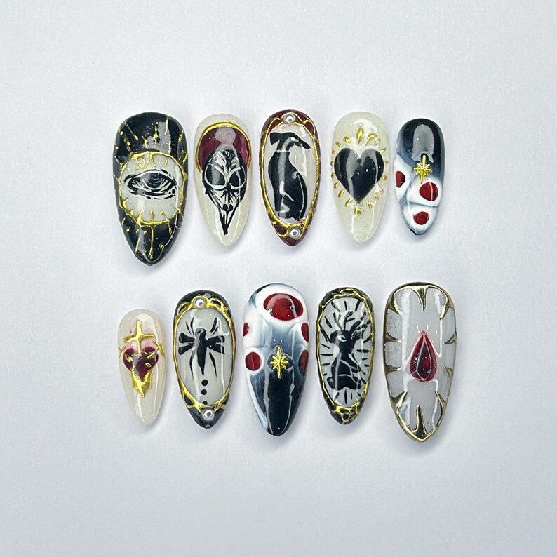 May include: A collection of ten almond-shaped false nails with gothic designs. Each nail showcases unique artwork, including an eye, skull, heart, and spider, with gold detailing. The nails are painted in black, white, red, and grey.