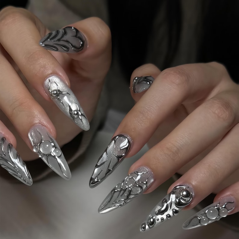 May include: Silver and gray stiletto nails with intricate designs. The nails feature a variety of embellishments, including metallic accents, geometric patterns, and 3D elements. The overall aesthetic is futuristic and edgy.