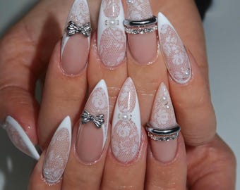 May include: A set of long, pointed artificial nails. The nails feature a white and nude color scheme with lace patterns, silver bows, and pearl accents. Some nails have silver rings. The overall design is elegant and feminine.