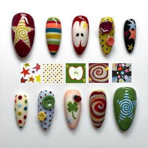 Puede incluir: Una colección de uñas artificiales pintadas con varios diseños. Los diseños incluyen estrellas, rayas, lunares, una manzana, un trébol de cuatro hojas y espirales. Los colores incluyen rojo, amarillo, verde, azul y naranja. Algunas uñas tienen botones o una mariquita.