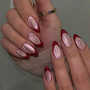 May include: Close-up of manicured nails with a red French tip and a shimmering, light pink base. The nails are oval-shaped and long. The image also shows multiple rings with colorful gemstones and gold bands.