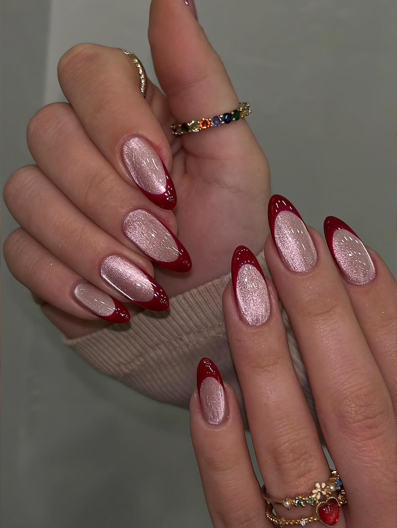 May include: Close-up of manicured nails with a red French tip and a shimmering, light pink base. The nails are oval-shaped and long. The image also shows multiple rings with colorful gemstones and gold bands.