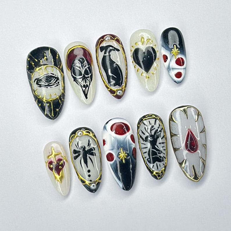 May include: A collection of ten almond-shaped artificial nails, each hand-painted with gothic designs. The designs include an eye, a skull, a raven, a heart, and a spider, with gold and red detailing. The nails are displayed on a white surface.