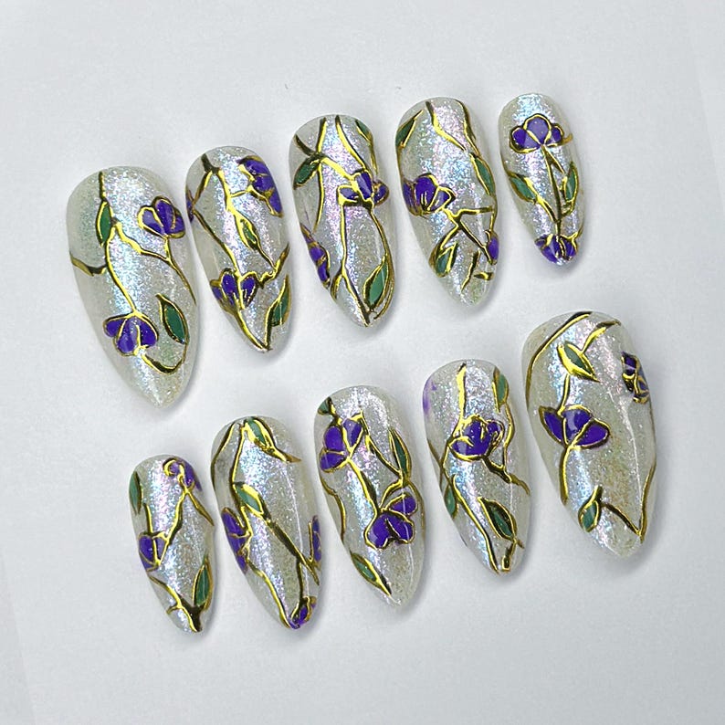 May include: Set of ten artificial nails with a pearlescent finish. Each nail is adorned with a floral design featuring purple flowers, green leaves, and gold-colored stems. The nails are oval-shaped and arranged on a white surface.