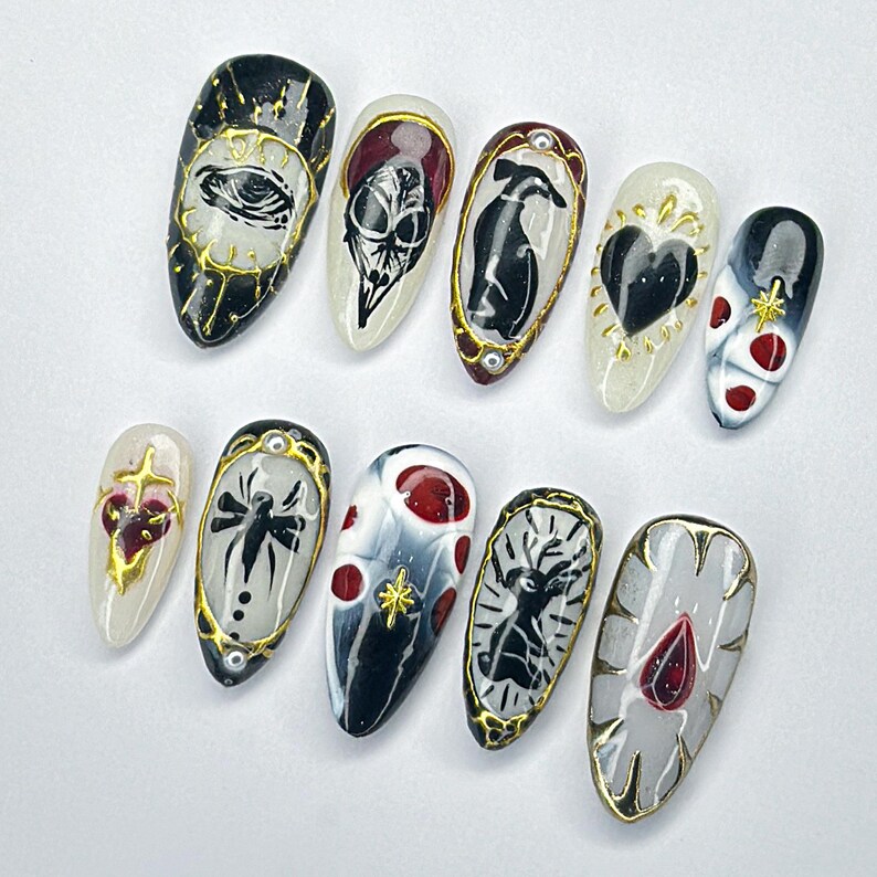 May include: A collection of ten artificial nails, each adorned with gothic-themed artwork. Designs include an eye, skull, heart, spider, and deer, with gold and red detailing. The almond-shaped nails are displayed against a white backdrop.