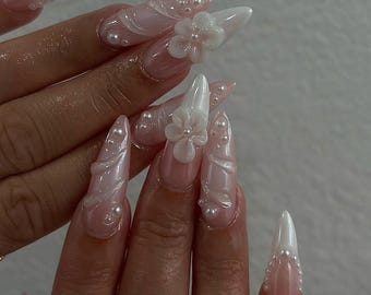 May include: Long, pointed acrylic nails with a light pink base and white tips. The nails are adorned with white 3D flower accents and small pearl embellishments, creating a delicate and elegant look. The nails are a fashionable accessory.