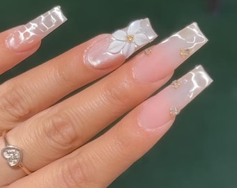 May include: Long square-shaped artificial nails featuring a soft pink base with a glossy finish. The design includes three-dimensional white floral accents with gold centers and small clear rhinestones placed on the nail beds. A silver heart ring.
