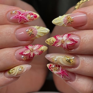 May include: A set of almond-shaped, professionally manicured nails. Each nail features a unique design with pink, yellow, and white floral and starfish accents. Gold embellishments and small pearls add detail to the nail art.