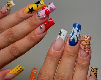 Puede incluir: Un conjunto de uñas acrílicas largas y cuadradas con varios diseños. Las uñas presentan una gama de colores, incluyendo amarillo con estrellas negras, rosa con corazones rojos y azul con adornos blancos. Algunas uñas tienen tachuelas plateadas y detalles dorados.