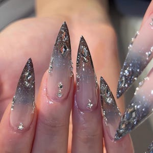 May include: Close-up of long, pointed fingernails with a gradient design. The nails transition from a clear base to a dark, glittery tip. Embellished with silver rhinestones, the nails have a glamorous, elegant appearance.