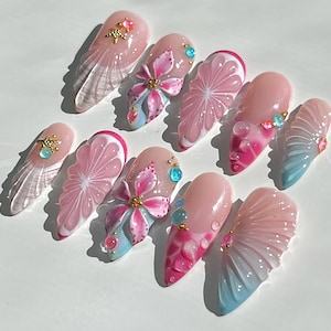 May include: A collection of ten artificial nails, each with a unique design. The nails are primarily light pink, adorned with floral patterns, gemstones, and gradient effects in pink and blue. Some nails feature a clear, textured design.