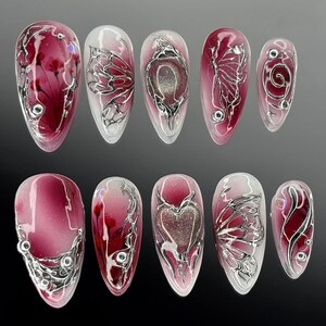 Puede incluir: Un conjunto de diez u&ntilde;as artificiales con base transparente y diversos dise&ntilde;os. Las u&ntilde;as presentan un degradado de borgo&ntilde;a y blanco, con acentos met&aacute;licos plateados en formas florales, de mariposas y de coraz&oacute;n. Algunas u&ntilde;as tienen peque&ntilde;os adornos plateados.