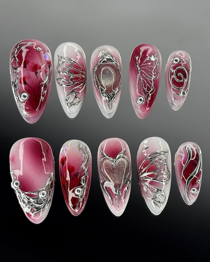 Puede incluir: Un conjunto de diez u&ntilde;as artificiales con base transparente y diversos dise&ntilde;os. Las u&ntilde;as presentan un degradado de borgo&ntilde;a y blanco, con acentos met&aacute;licos plateados en formas florales, de mariposas y de coraz&oacute;n. Algunas u&ntilde;as tienen peque&ntilde;os adornos plateados.