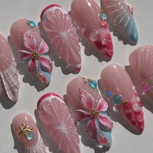 May include: A collection of ten artificial nails with a pale pink base. Each nail showcases detailed designs, such as pink flowers, citrus fruit slices, and iridescent accents. Some nails have blue and gold embellishments, creating a delicate and feminine look.