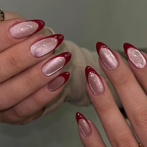 May include: Close-up of a hand with long, oval-shaped nails. The nails feature a French manicure design with a shimmering, light pink base and a deep red tip. A gold ring with floral and heart details is visible.