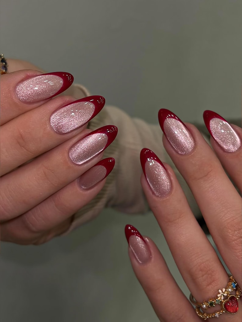 May include: Close-up of a hand with long, oval-shaped nails. The nails feature a French manicure design with a shimmering, light pink base and a deep red tip. A gold ring with floral and heart details is visible.